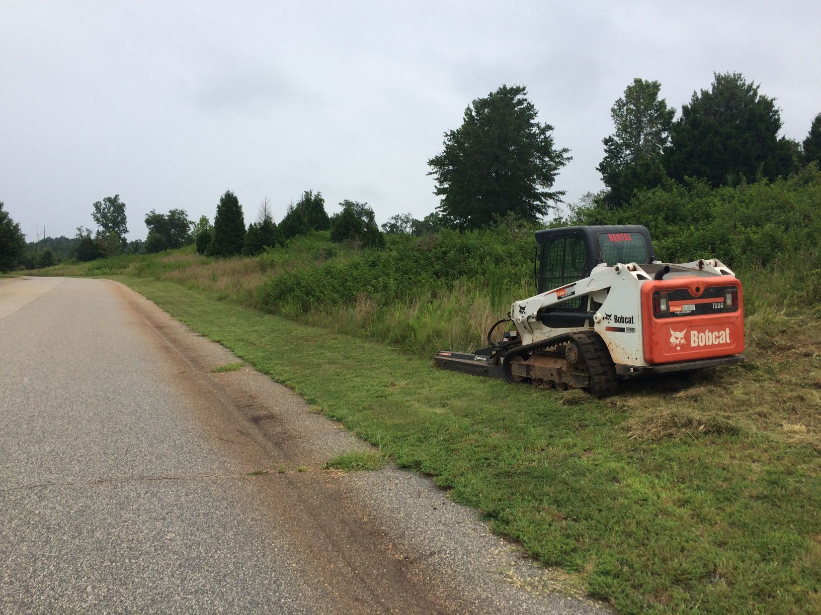 Bobcat mower cutting grass next to a road. Cloudy day, green grass, brown dirt.