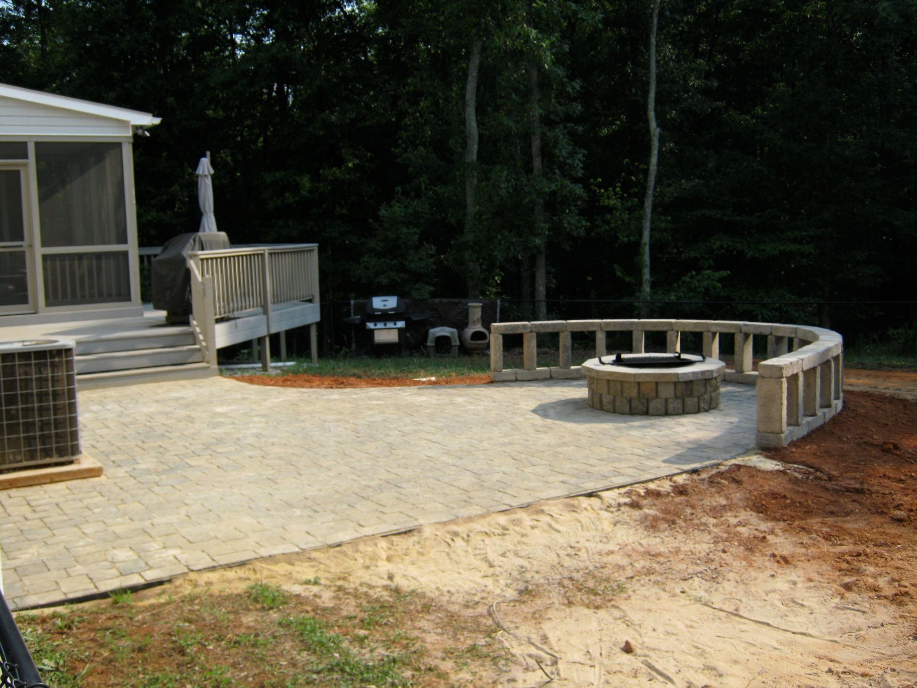 Patio with a fire pit, bordering wall, and a deck connected to a house in a wooded area.