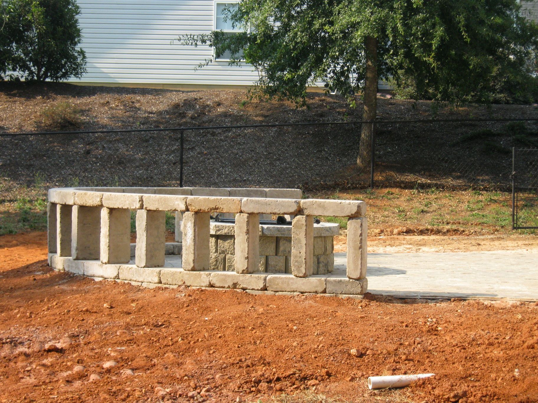 Stone structure in a dirt yard, with a black fence and a house in the background.