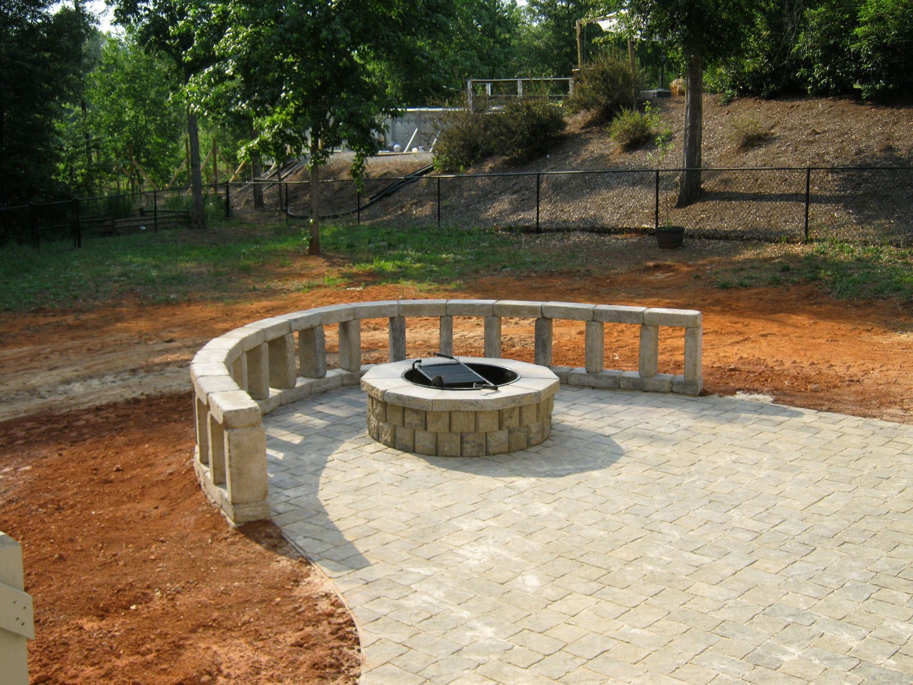 Curved stone wall surrounds a brick patio with a circular fire pit. Brown dirt surrounds the patio.