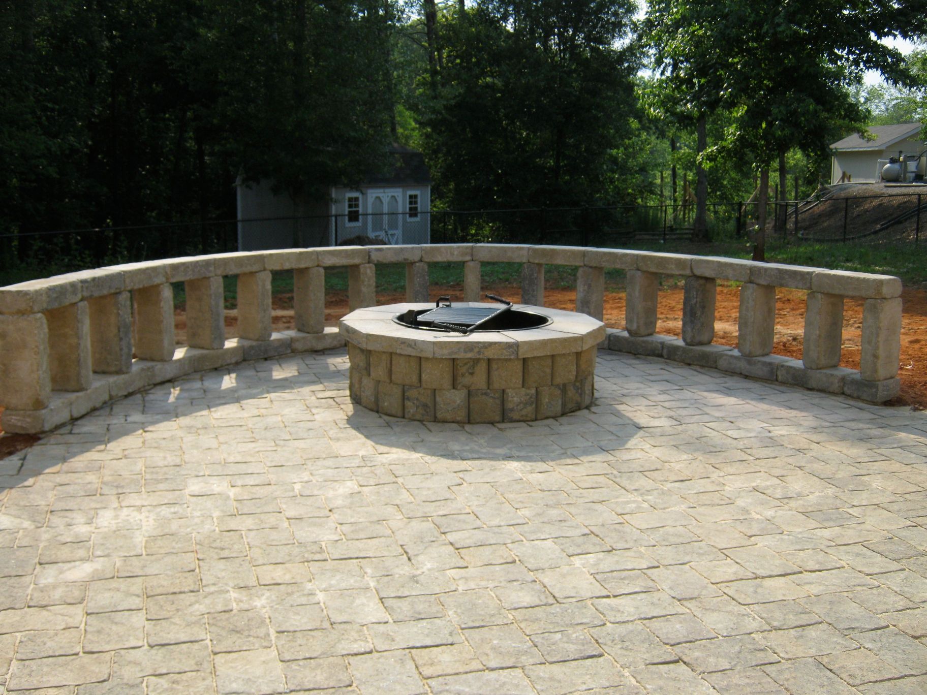 A curved stone patio with a circular fire pit and a low wall, in a backyard setting.
