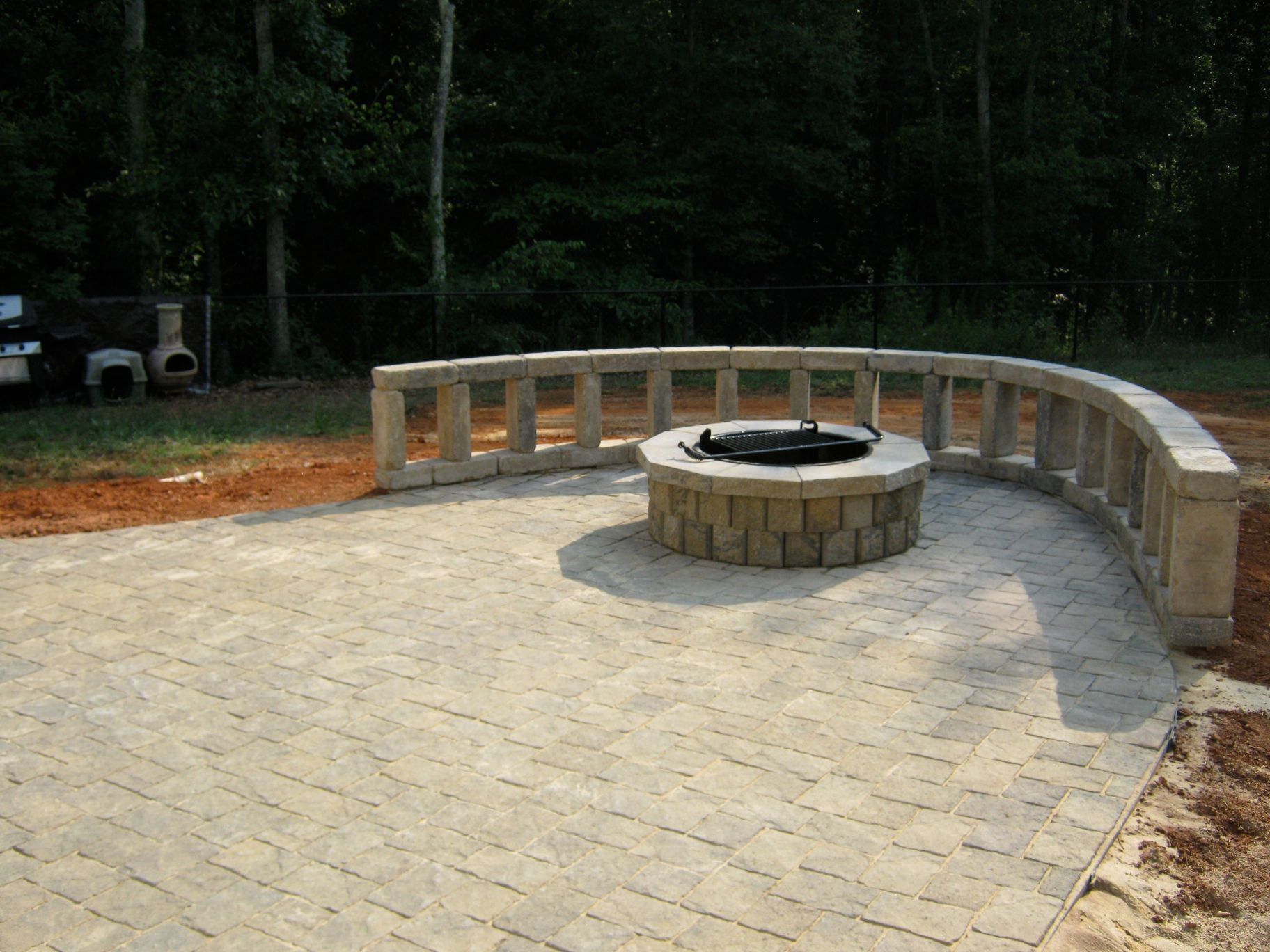 A circular patio with a stone fire pit and curved stone seating, surrounded by trees.