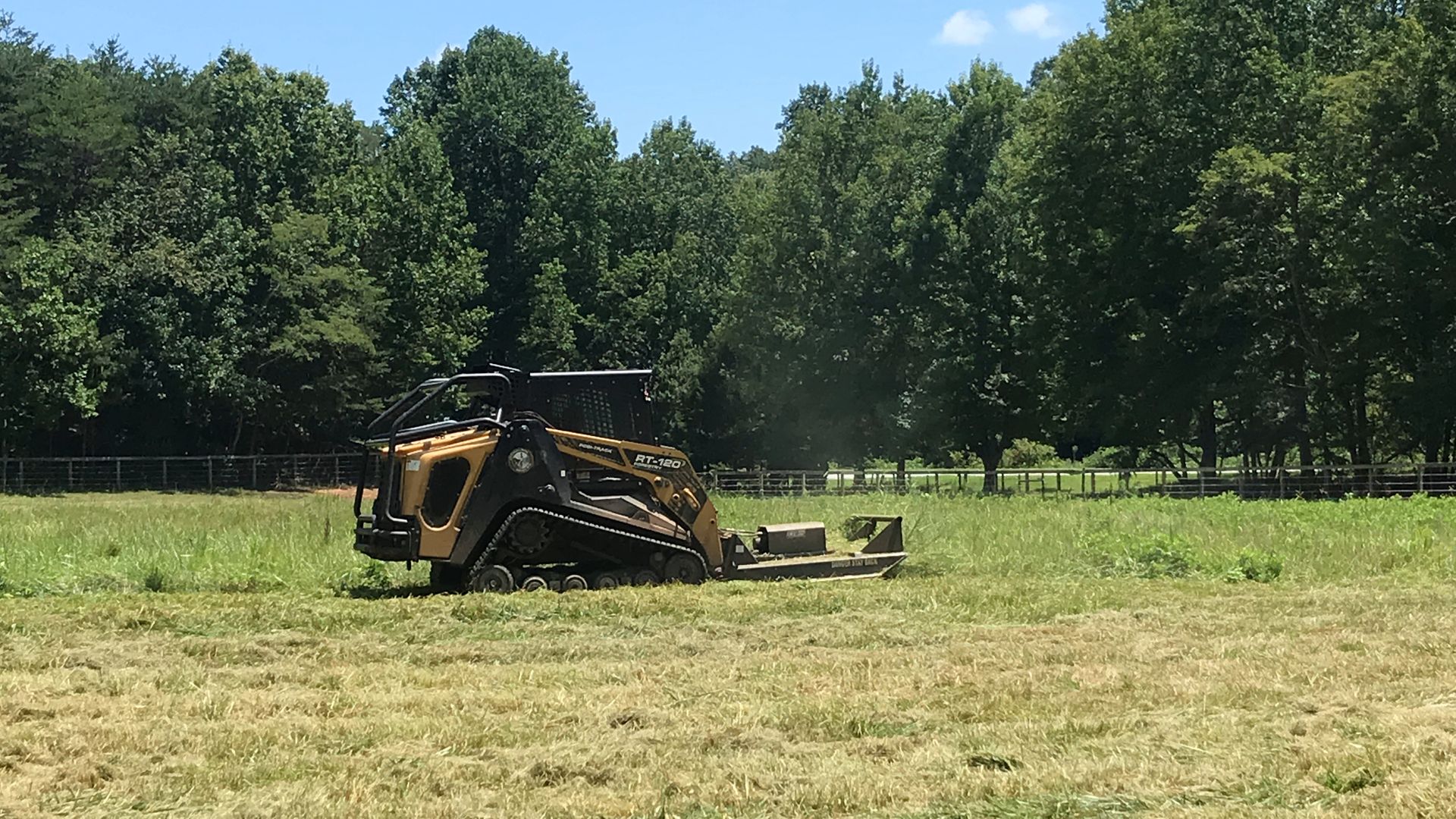 Yellow and black skid steer mower cutting grass in a field, trees in background.