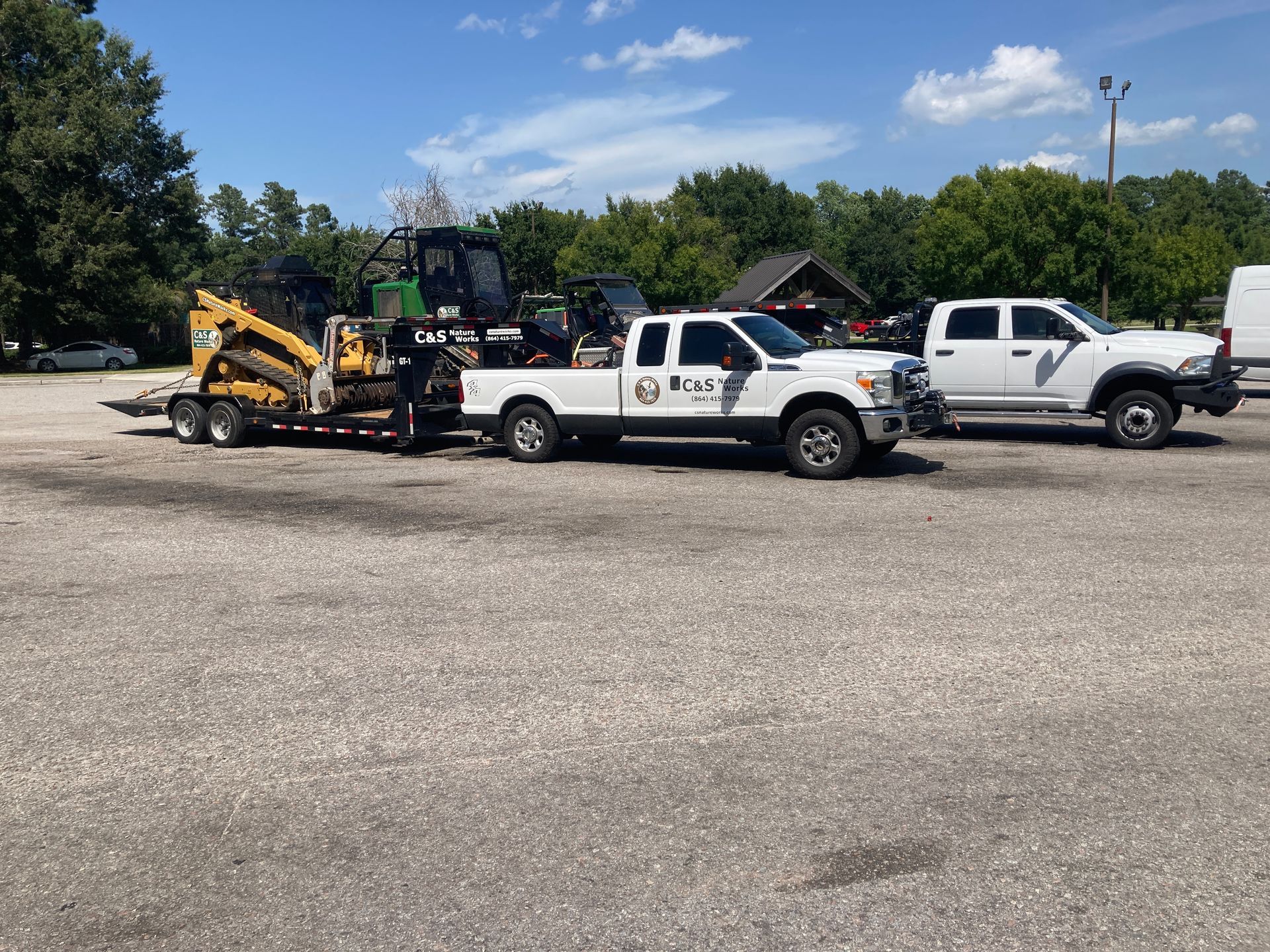 Two trucks, one towing a trailer with construction equipment, parked on a gravel lot on a sunny day.