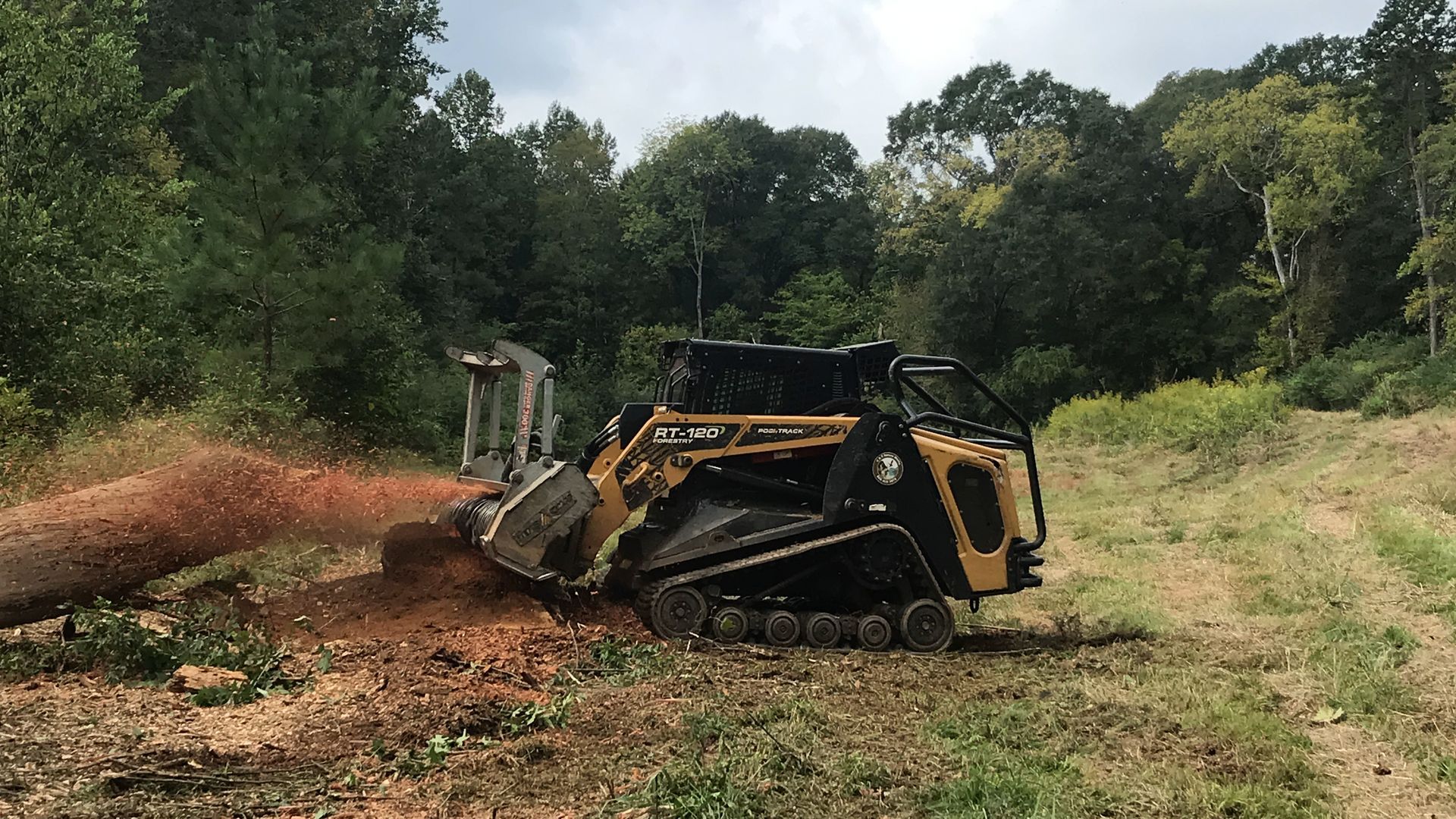 A small, yellow track loader clearing land, kicking up dirt, surrounded by trees.