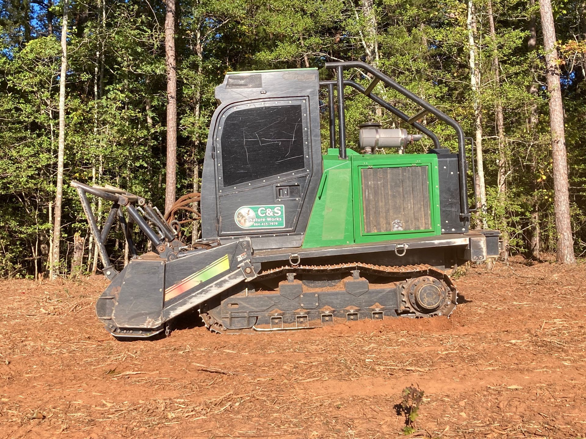 A green and black forestry mulcher on tracks in a wooded area.