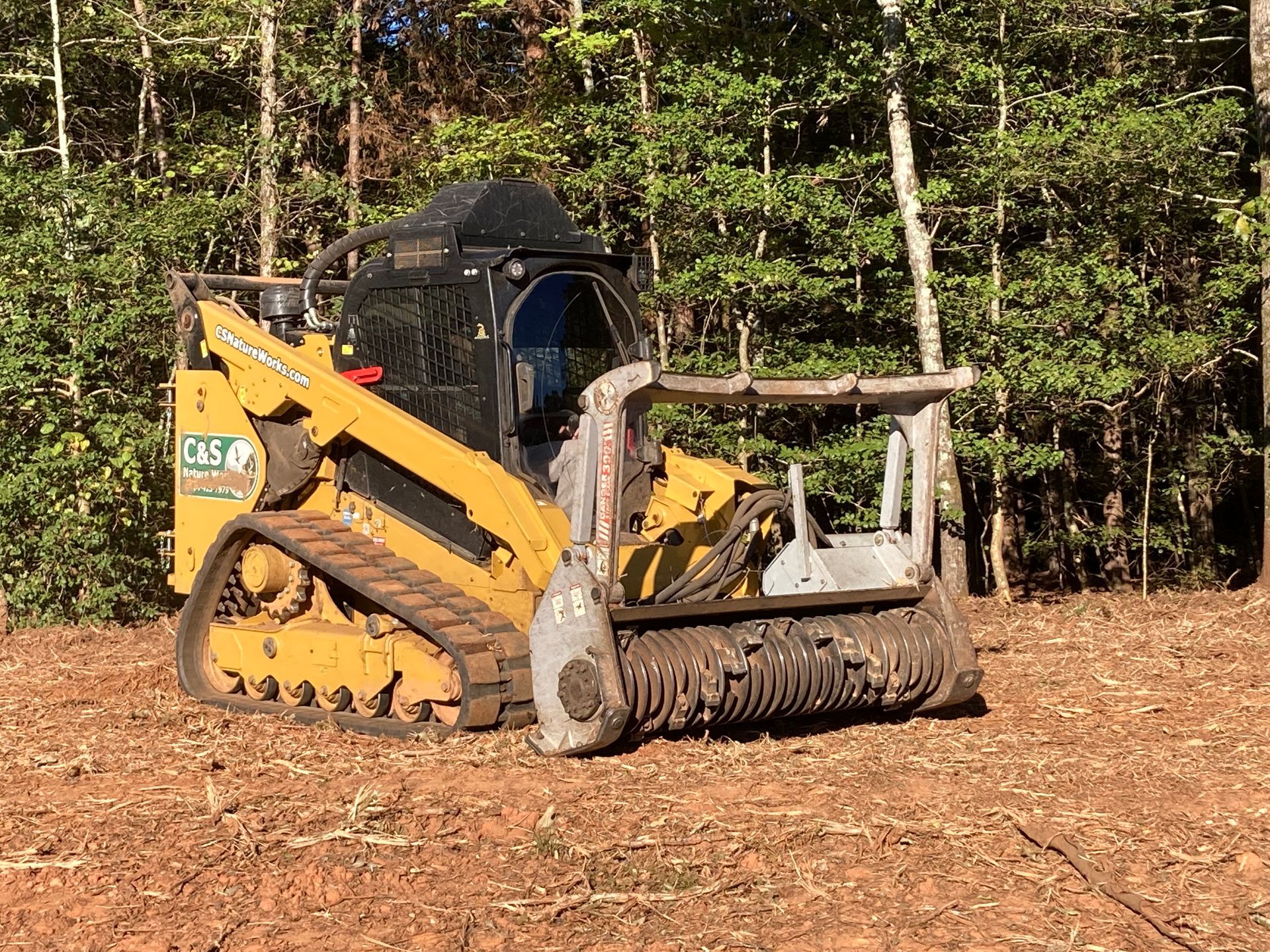 Yellow skid steer with brush cutter mulching land, trees in background.