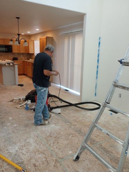 A person using a vacuum on a construction site with exposed subflooring, a ladder, and a kitchen visible in the background.