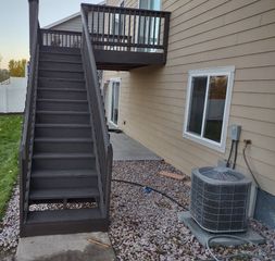 Dark wooden stairs lead up to a balcony on the side of a beige house with an AC unit nearby on a gravel ground.