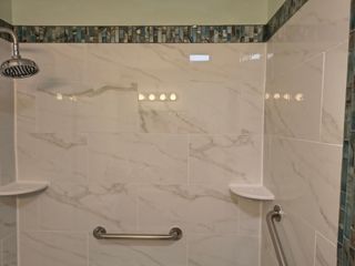 Shower stall featuring white marble-look tiles, a chrome shower head, grab bars, corner shelves, and a blue mosaic border.