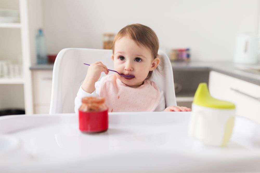 A baby is sitting in a high chair eating food with a spoon.