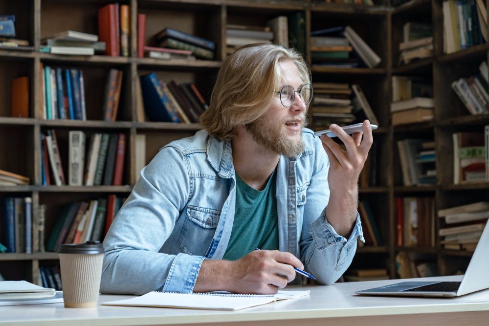 Blond man in glasses talking on phone, sitting at desk with books and laptop in library.