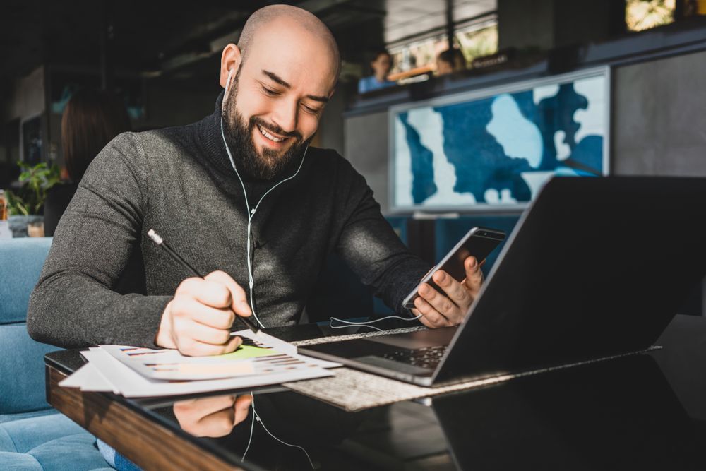 A smiling person with a beard sits at a table, holding a phone while taking notes on papers next to an open laptop.