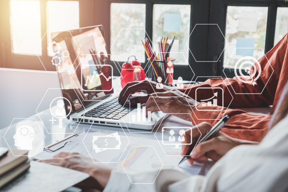 Two people working at a desk with a laptop and documents, digital icons overlay.