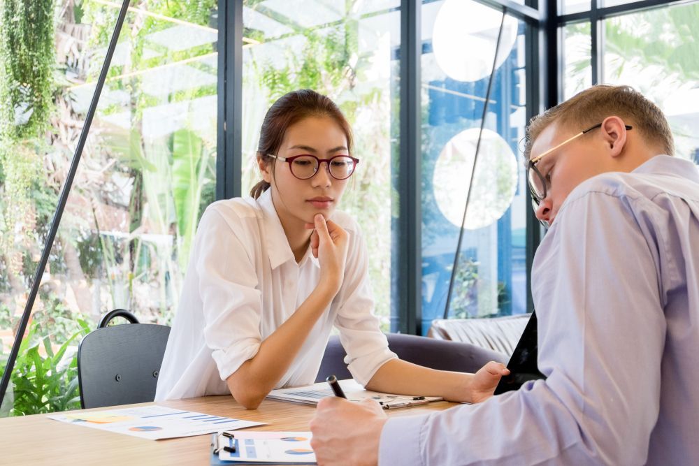 Woman in glasses, white shirt thoughtfully touches chin, looking at documents with a man in a light shirt and glasses at a table.