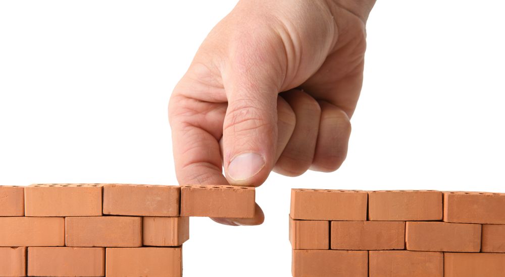 Hand placing a brick to complete a small wall of red bricks, white background.