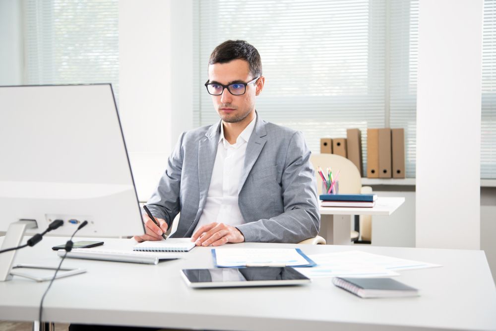 Man in glasses, blazer, working at a desk with computer and documents in an office.