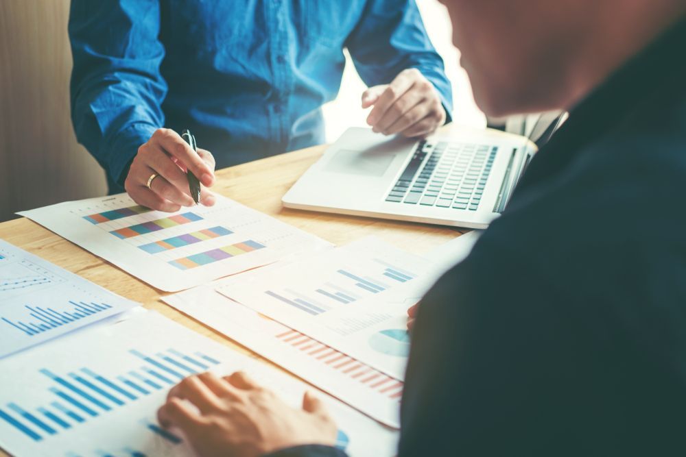 Two people reviewing data reports, graphs, and a laptop on a wooden desk.