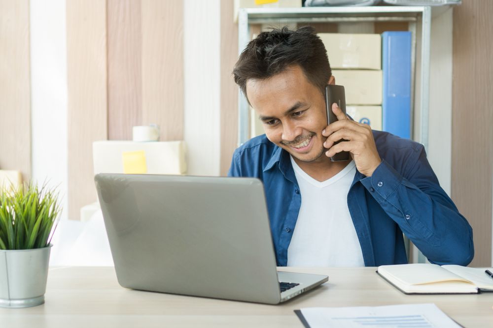 A man is sitting at a desk talking on a cell phone in front of a laptop computer.