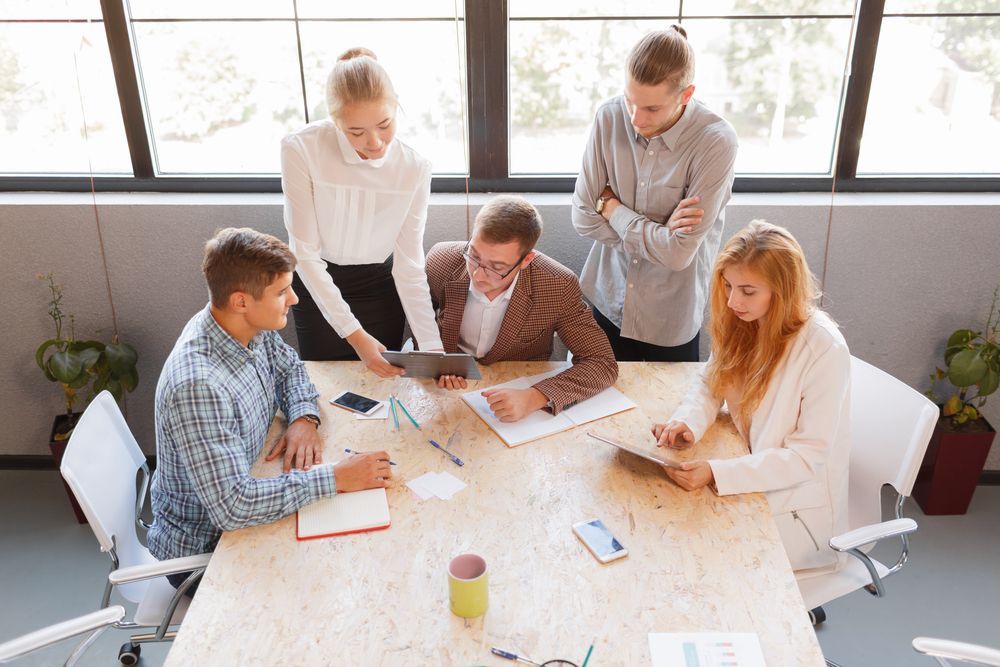 Group of people around a table looking at a tablet, discussing. Light-filled office setting.