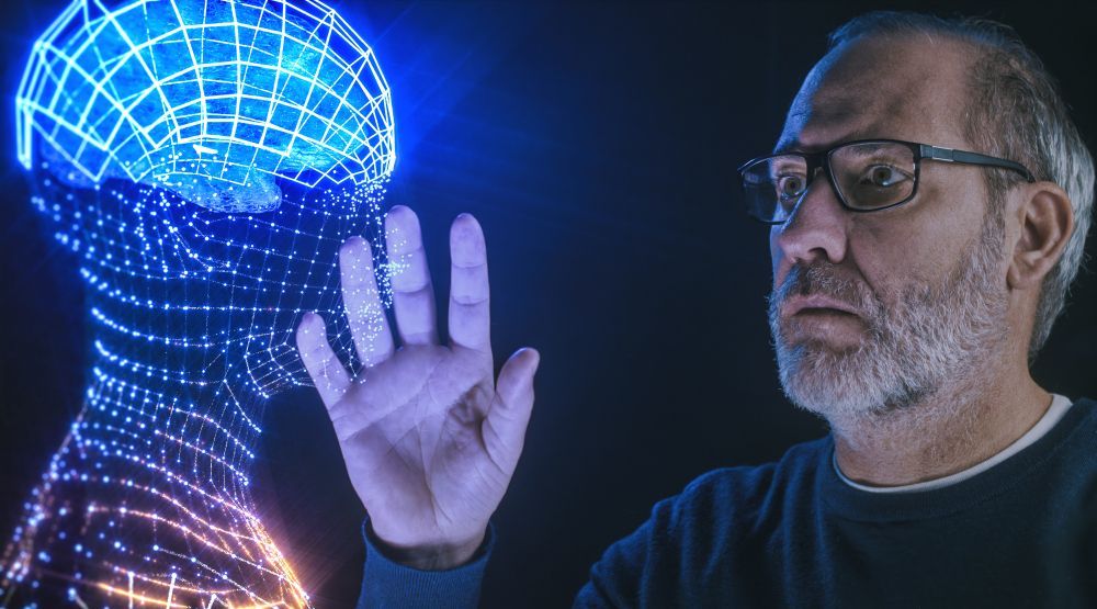 Man in glasses touches a glowing blue, wireframe head hologram with his hand. Dark background.