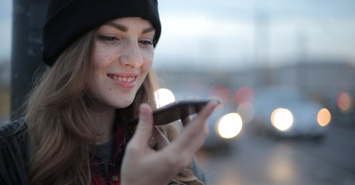 Woman with beanie smiling, holding phone, blurred street lights in background.