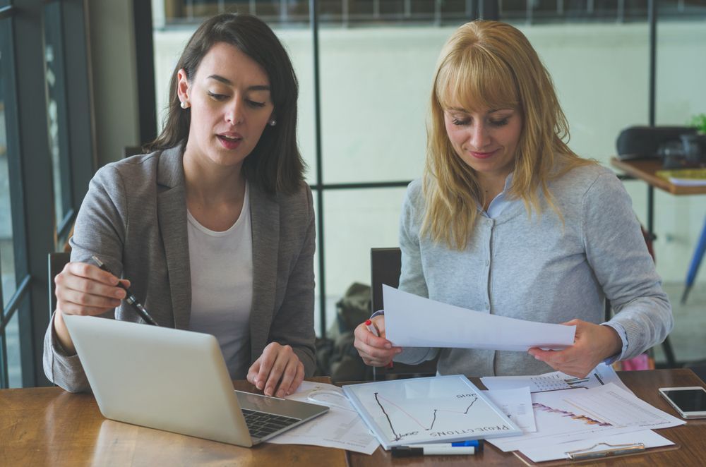 Two women are sitting at a table looking at papers and a laptop.