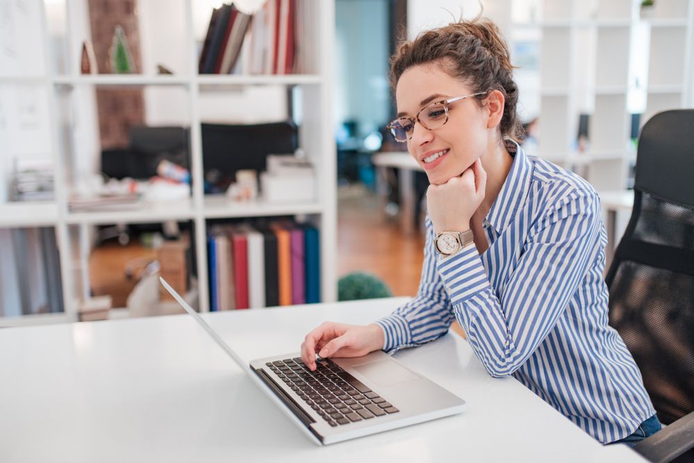 Woman in glasses at desk, using laptop, smiling in a bright office setting.