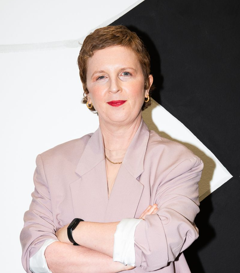 Woman in pink blazer and burgundy skirt, arms crossed, stands before black and white geometric wall.