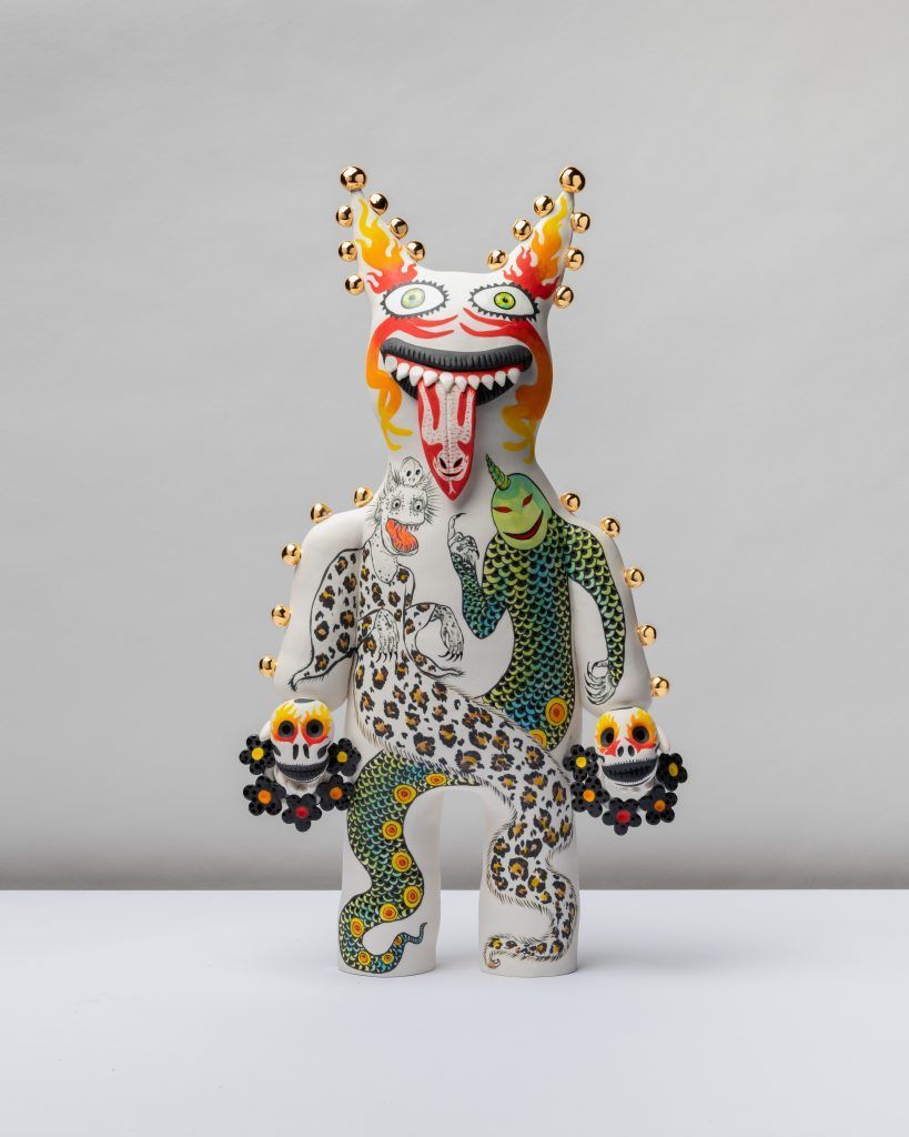 Whimsical creature sculpture. White body, decorated with flowers and faces, crowned with orange horns and a lolling tongue.