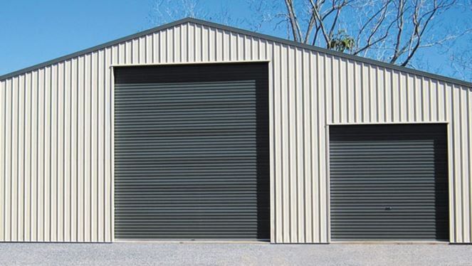Beige metal-clad shed with two dark gray roller doors, under a blue sky. Beige metal-clad shed with two dark gray roller doors, under a blue sky.