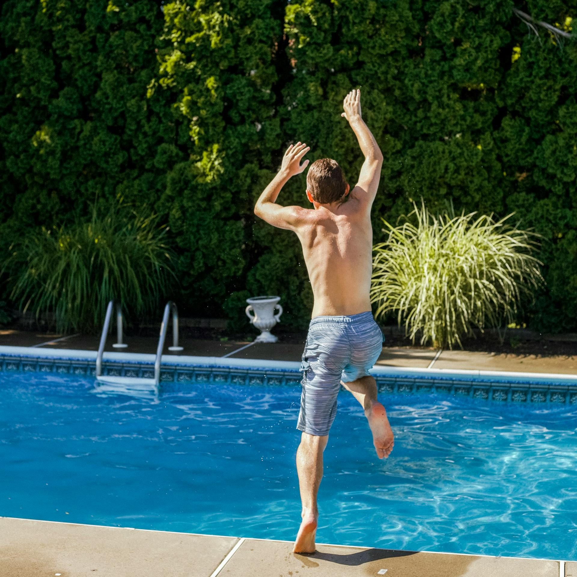 Man jumps into a pool with arms raised, blue water and shorts, green bushes in the background.
