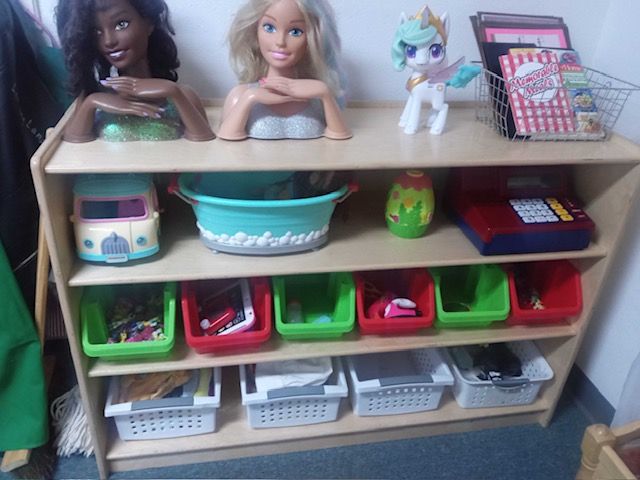Preschool classroom interior with tables, chairs, storage bins, and cabinets.