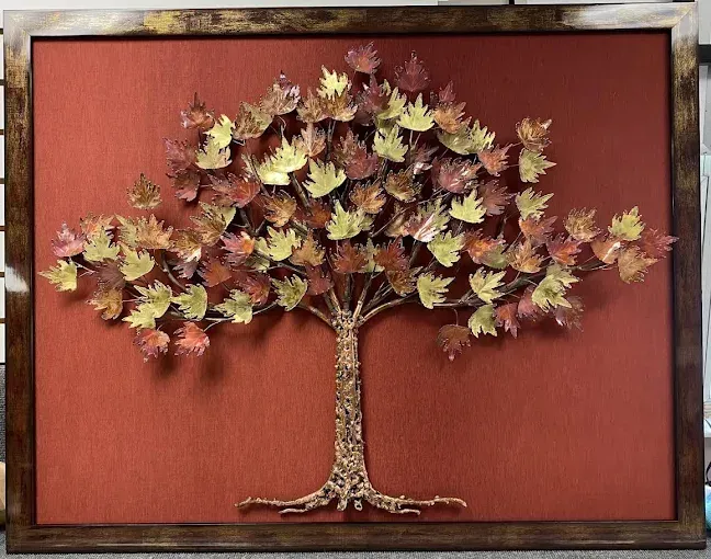 Metal art of a tree with leaves in autumn colors against a red textured background within a brown frame.