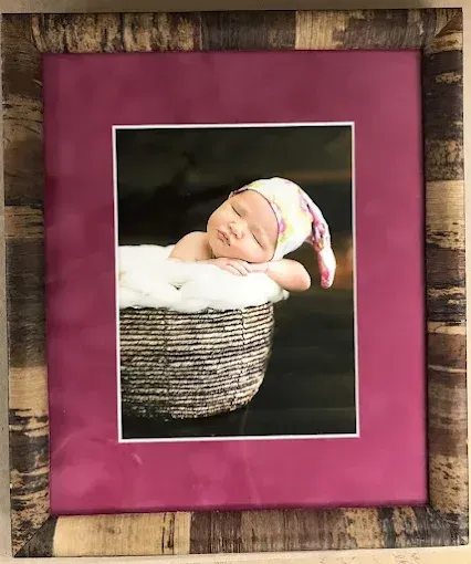 Newborn baby sleeping in a woven basket, wearing a hat, framed in pink and rustic wood.