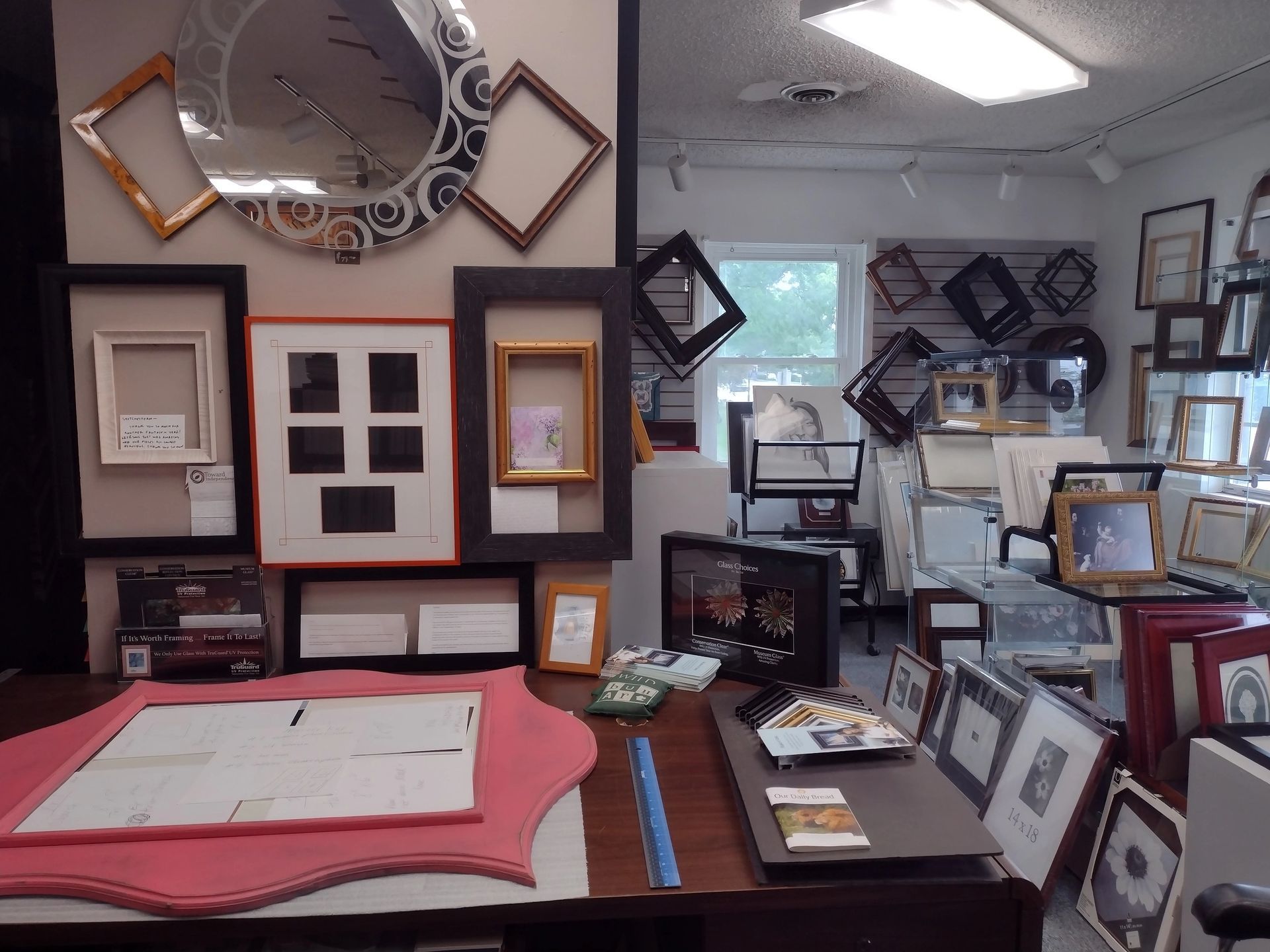 A custom framing shop displays framed art and mirrors on the wall. Frames in various sizes and styles are on display.