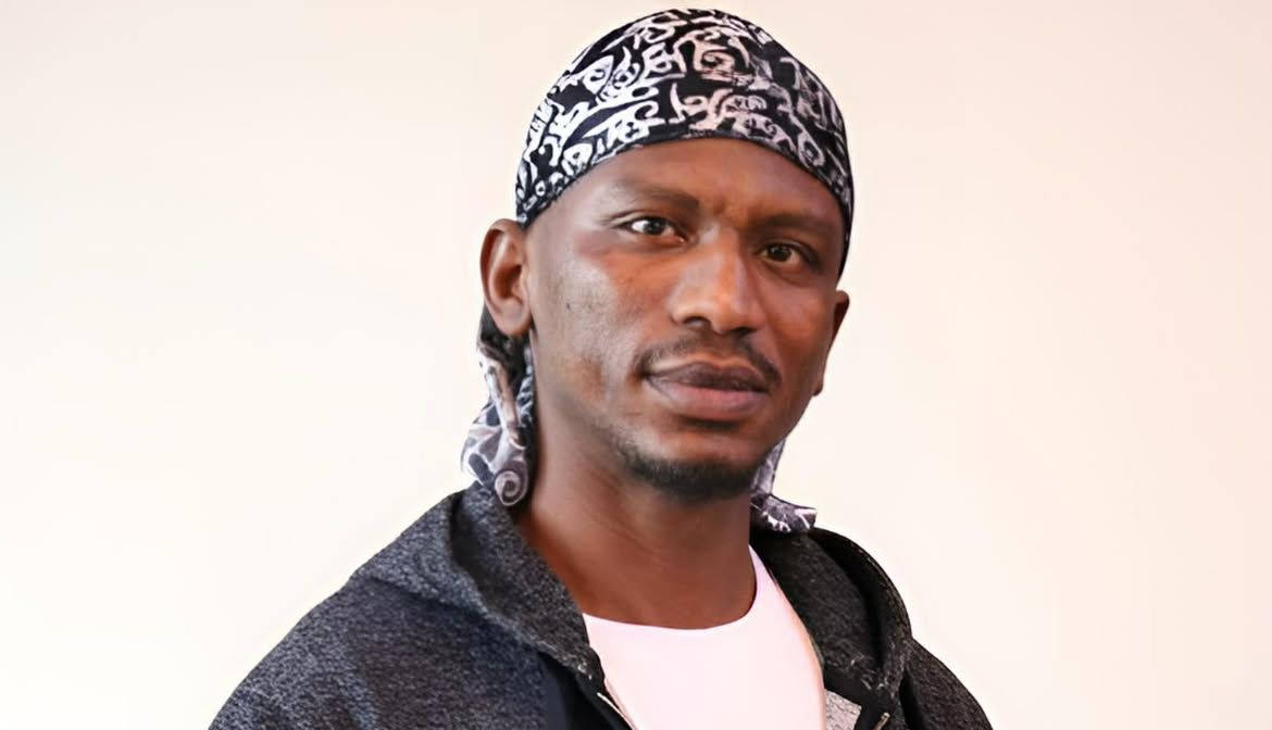 Man wearing a black and white patterned head covering and gray hooded sweatshirt, looking forward.