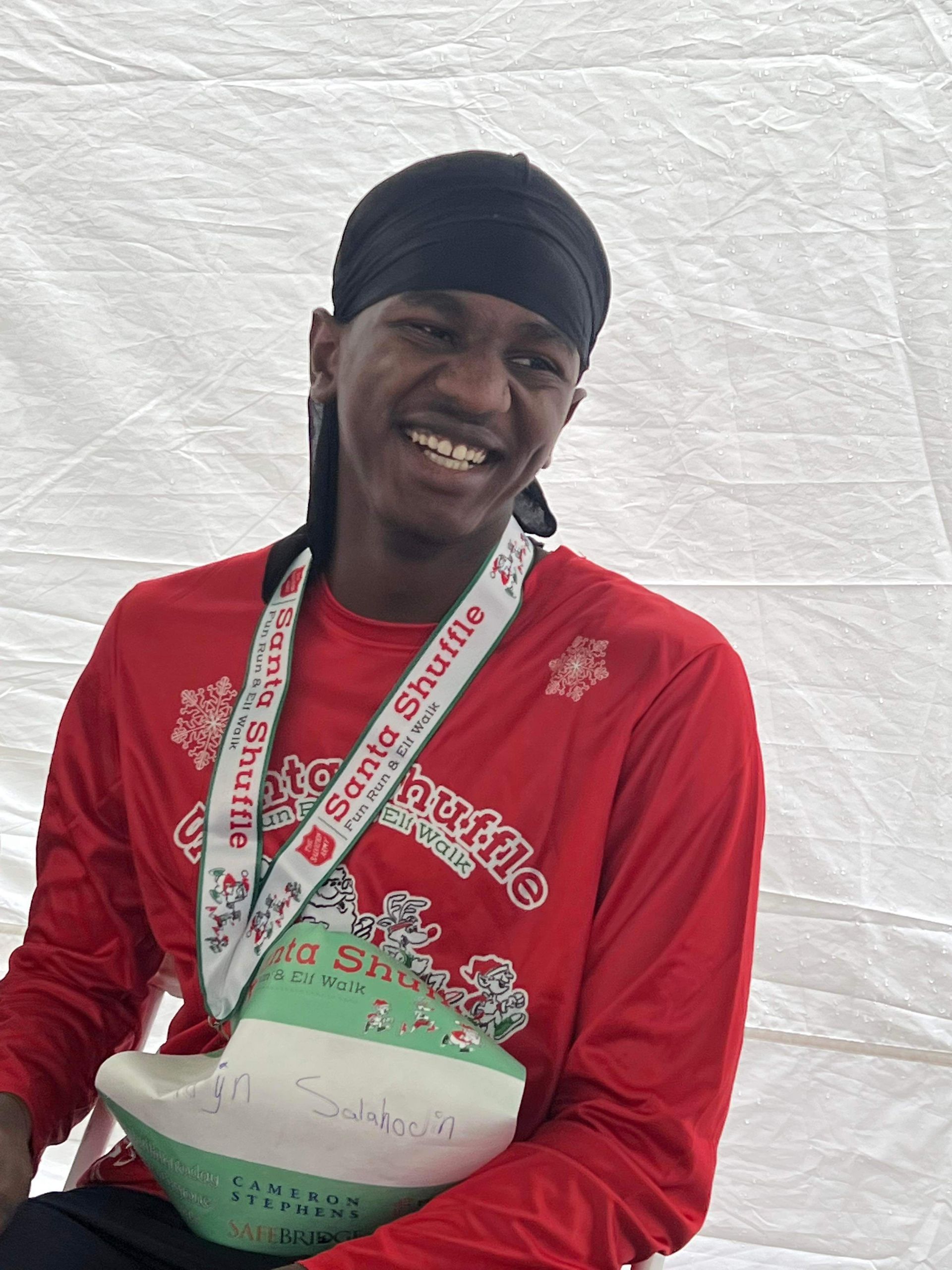 Person wearing a red shirt and black head covering smiles, wearing a Santa Shuttle medal.