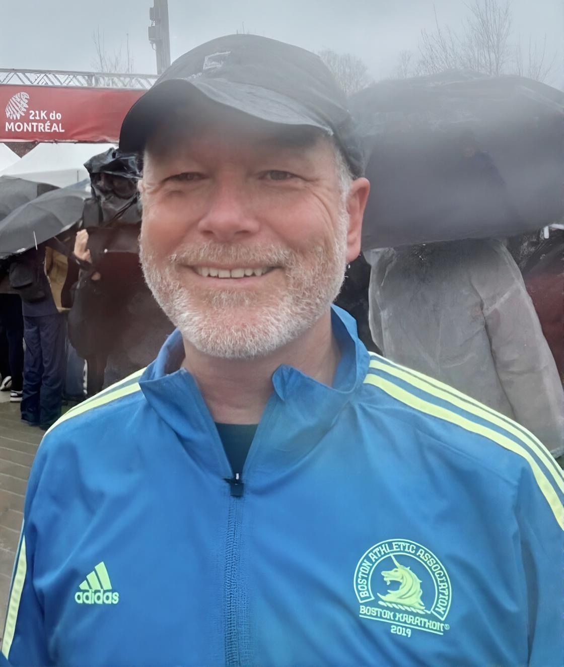 Man in blue jacket with Boston Marathon logo smiles at the finish line in the rain.
