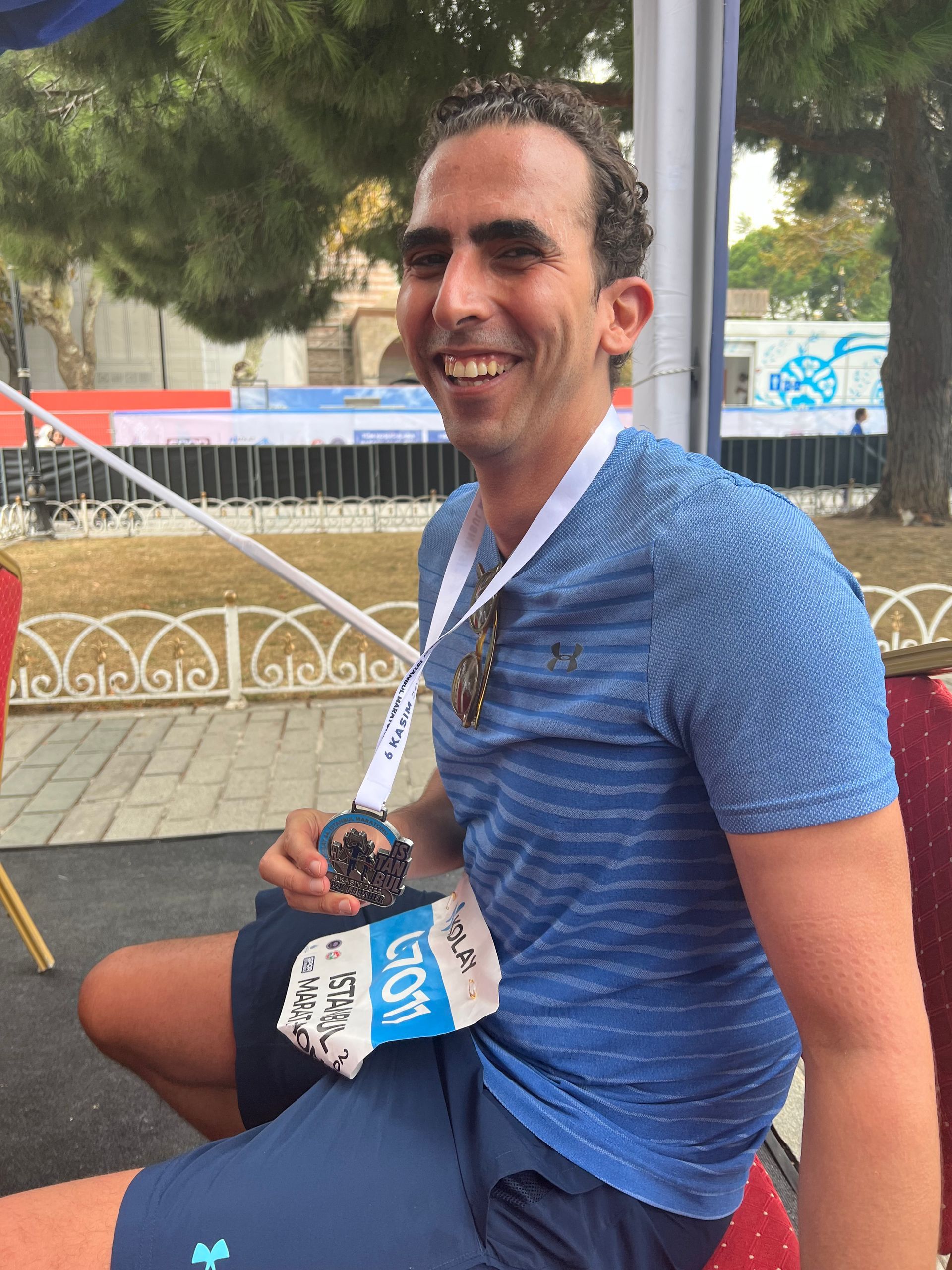 Man wearing a medal, smiling, sitting. Blue shirt and shorts. Outdoor setting with blurred background.