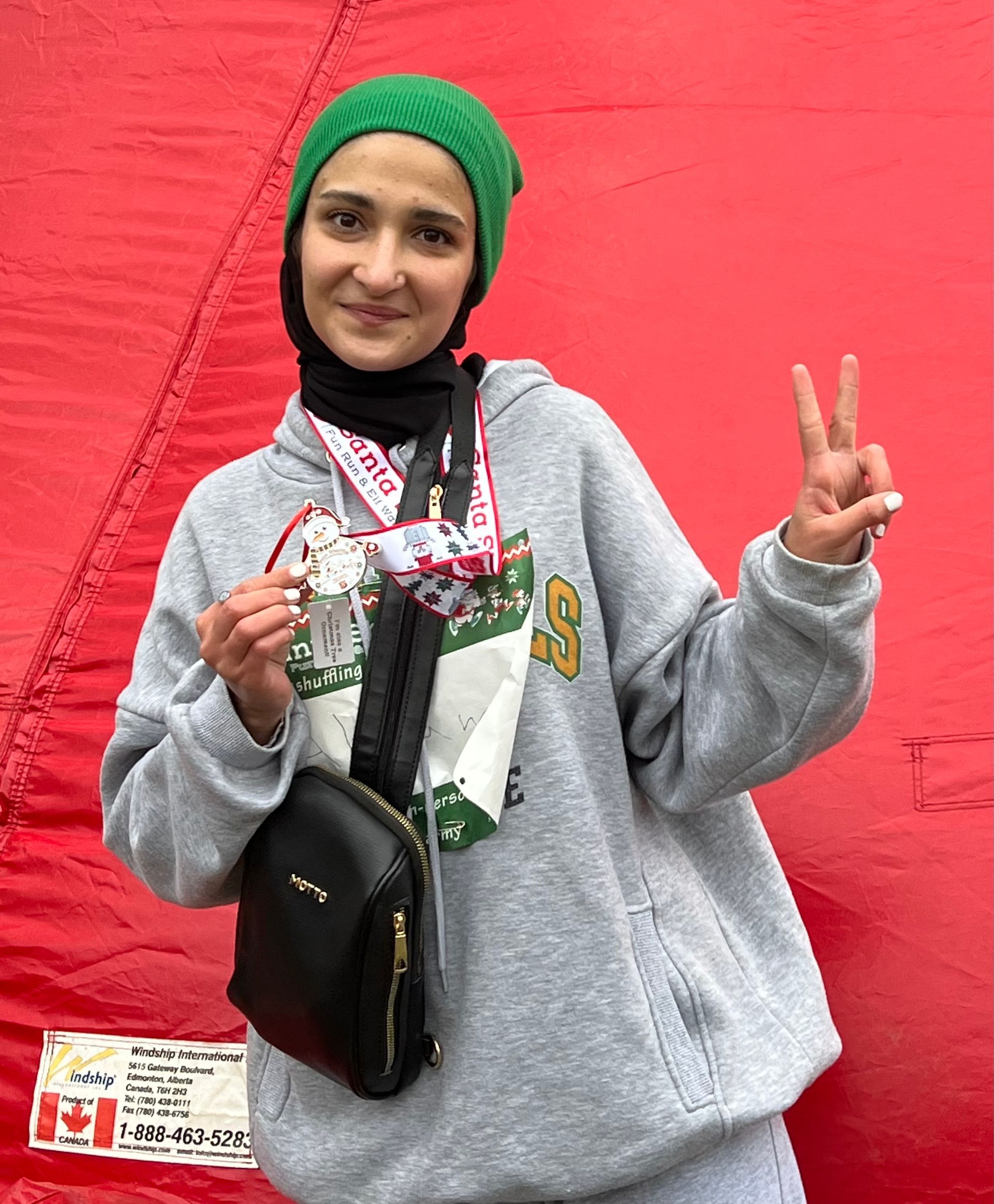 Woman in green hat and gray hoodie with a medal, giving the peace sign. Red background.