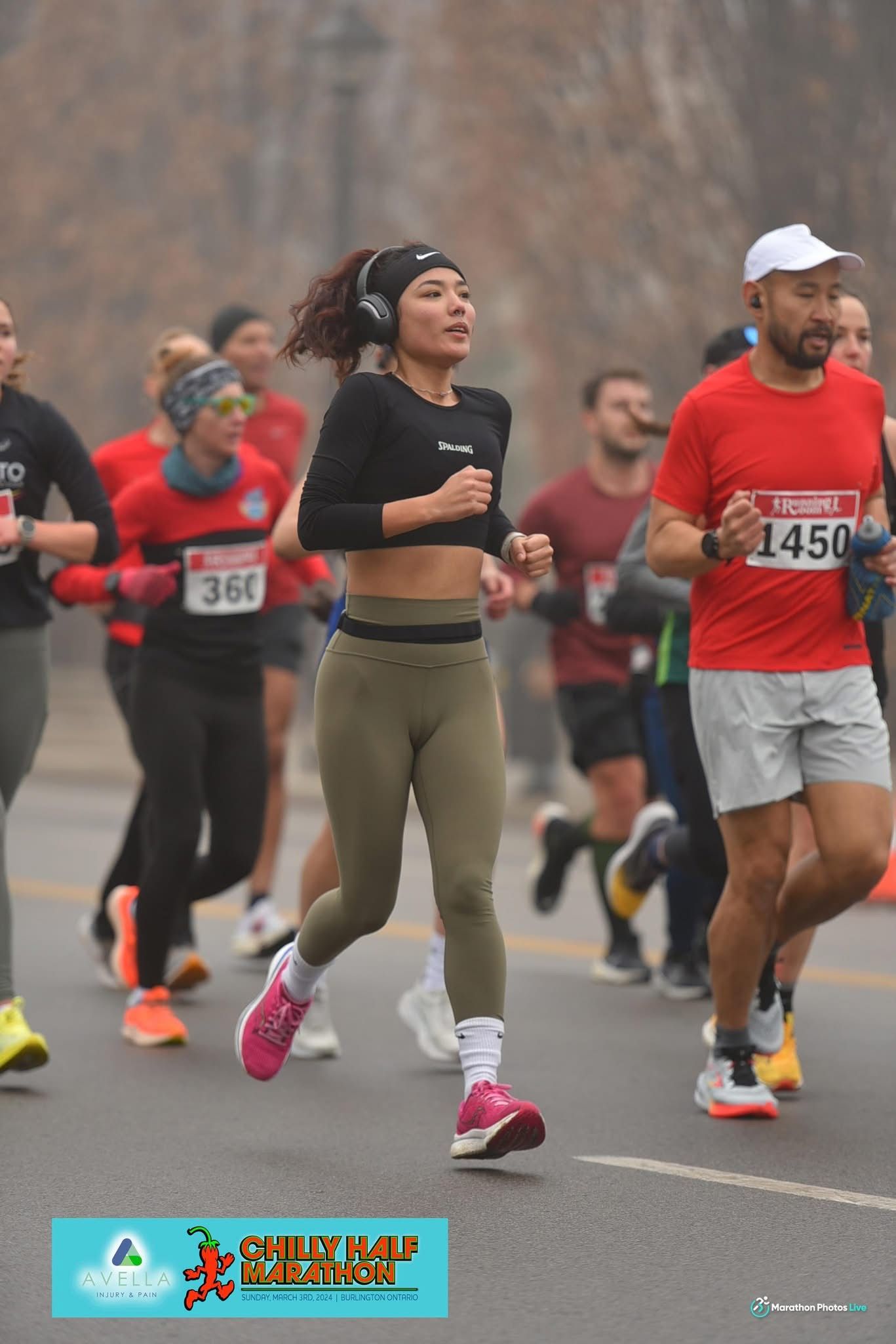 A woman running in a race, wearing a black top, green leggings, and pink shoes, on a road with other runners.