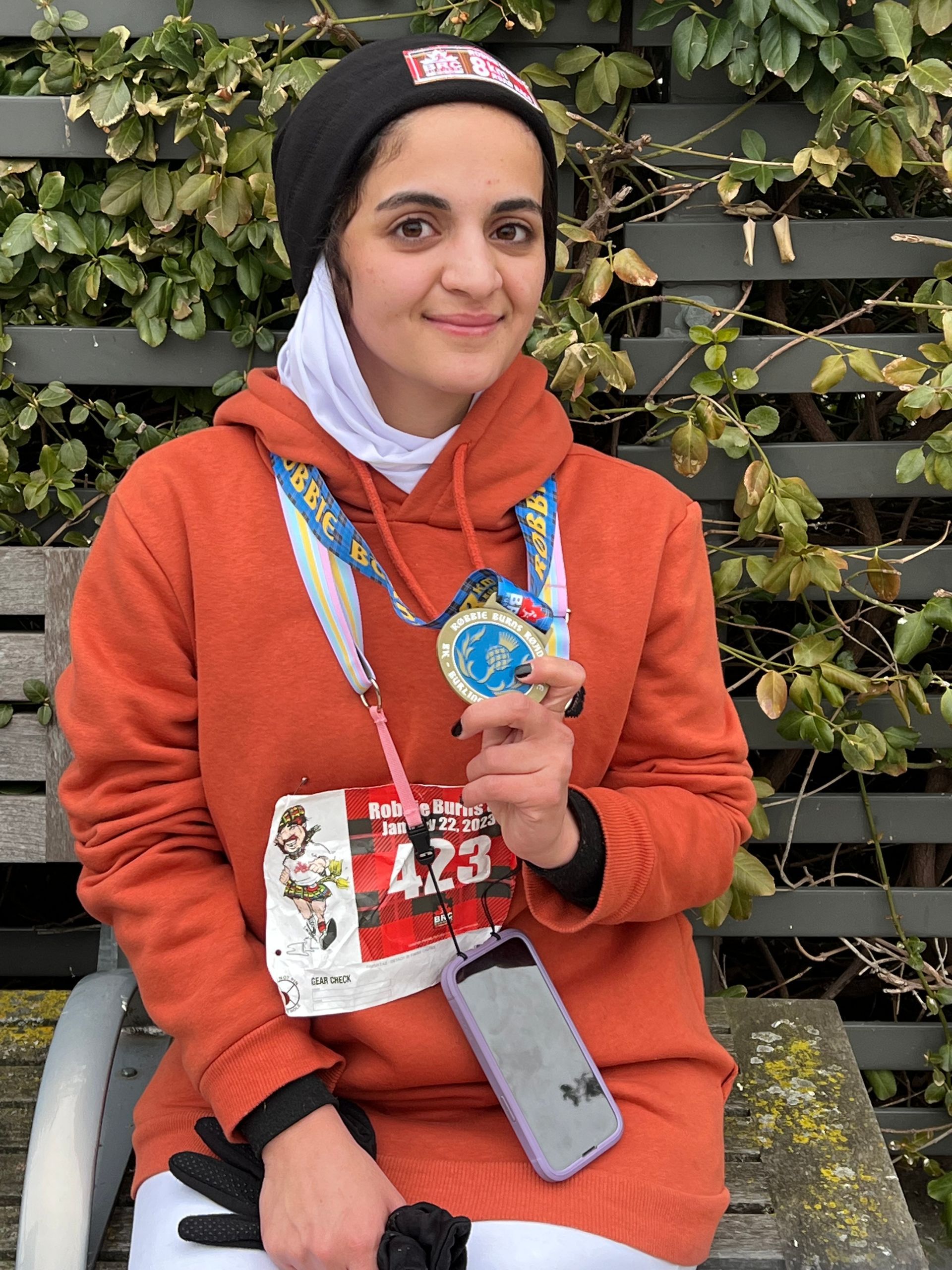 Woman in orange hoodie holds medal, race bib, and phone, sitting outside.