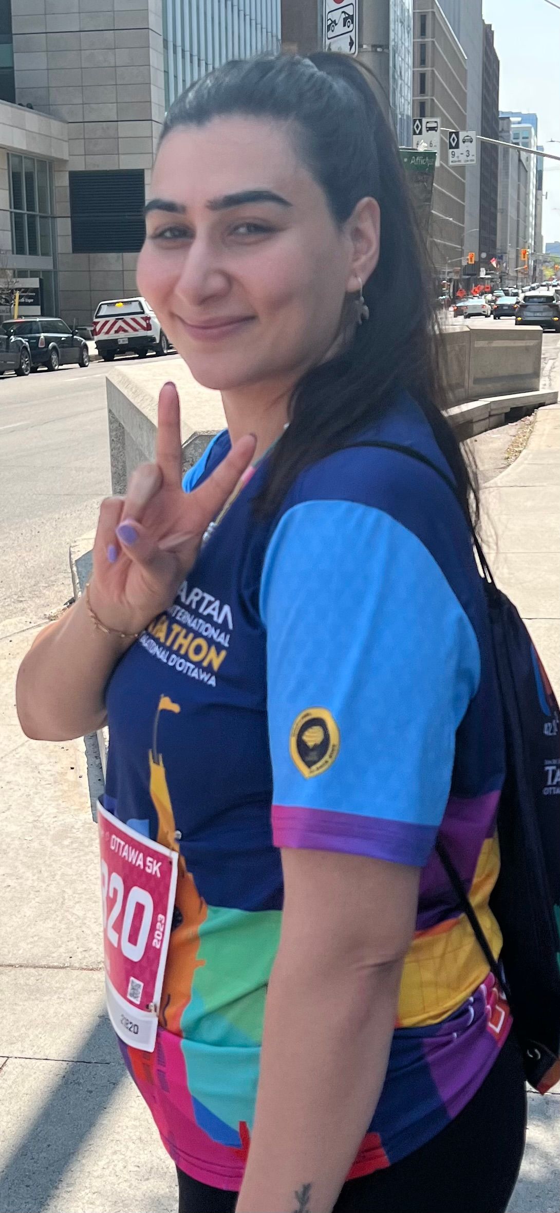 Woman giving peace sign during a marathon, wearing a colorful shirt and race bib. City background.