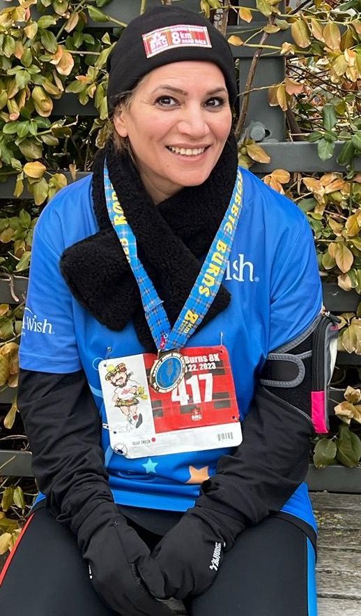 Woman in blue running shirt, medal, black hat and scarf, smiling.