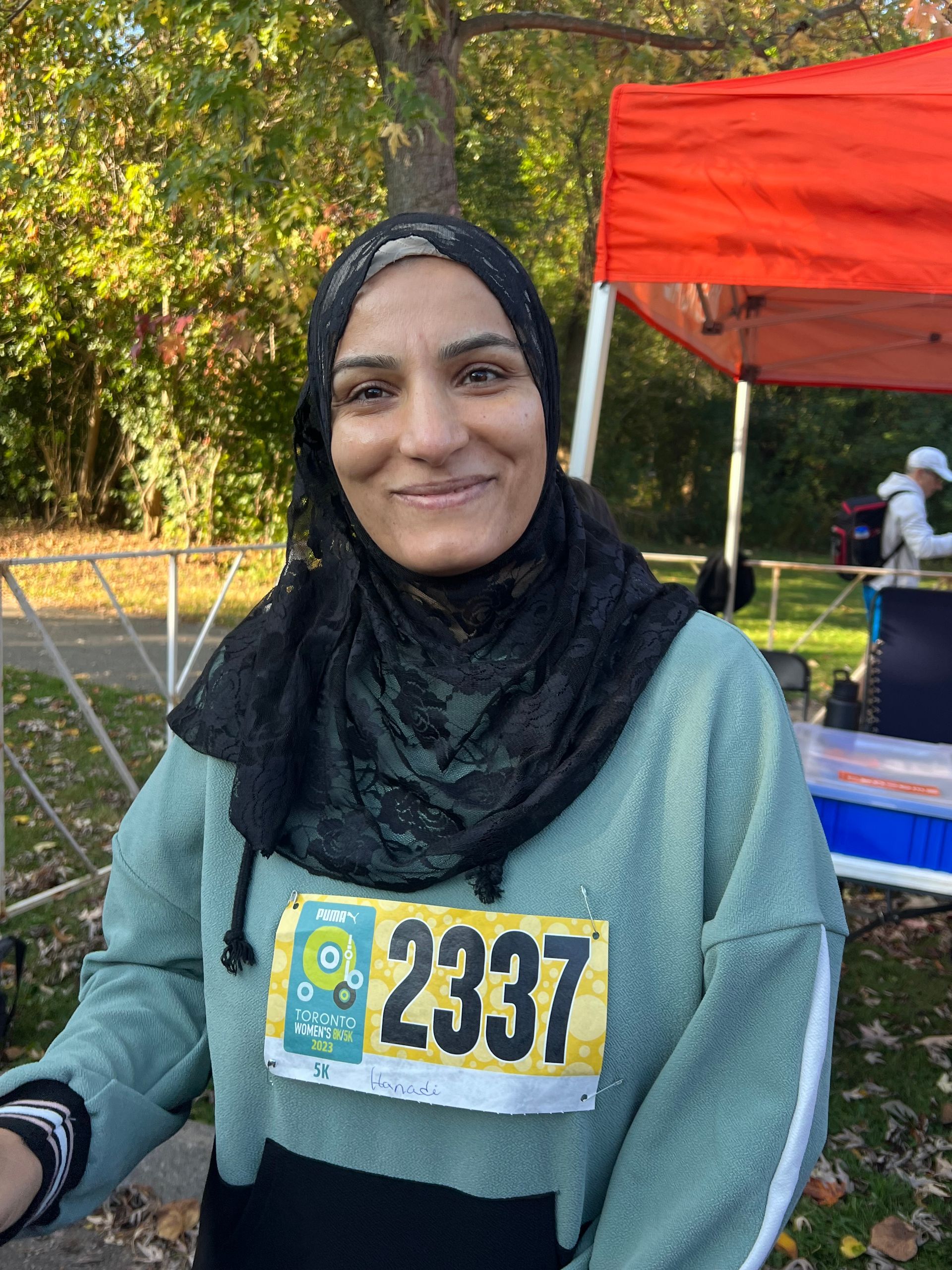 Woman in a black hijab and running bib with the number 2337 smiles, near an orange tent.