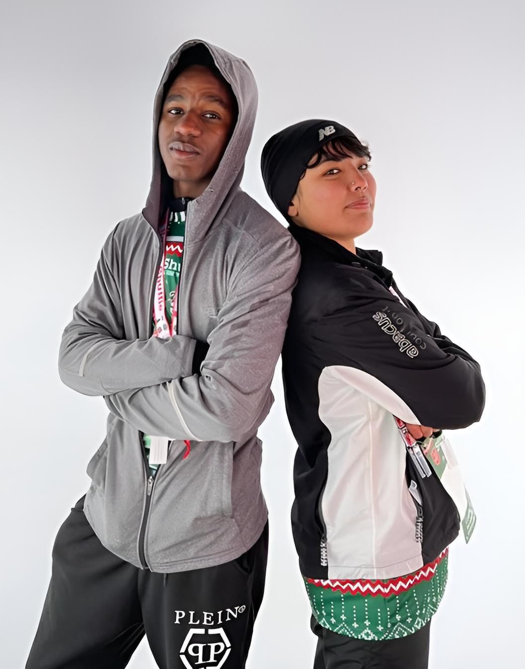 Two people in athletic wear stand back-to-back with arms crossed. One wears a grey hooded jacket, the other a black jacket and beanie.