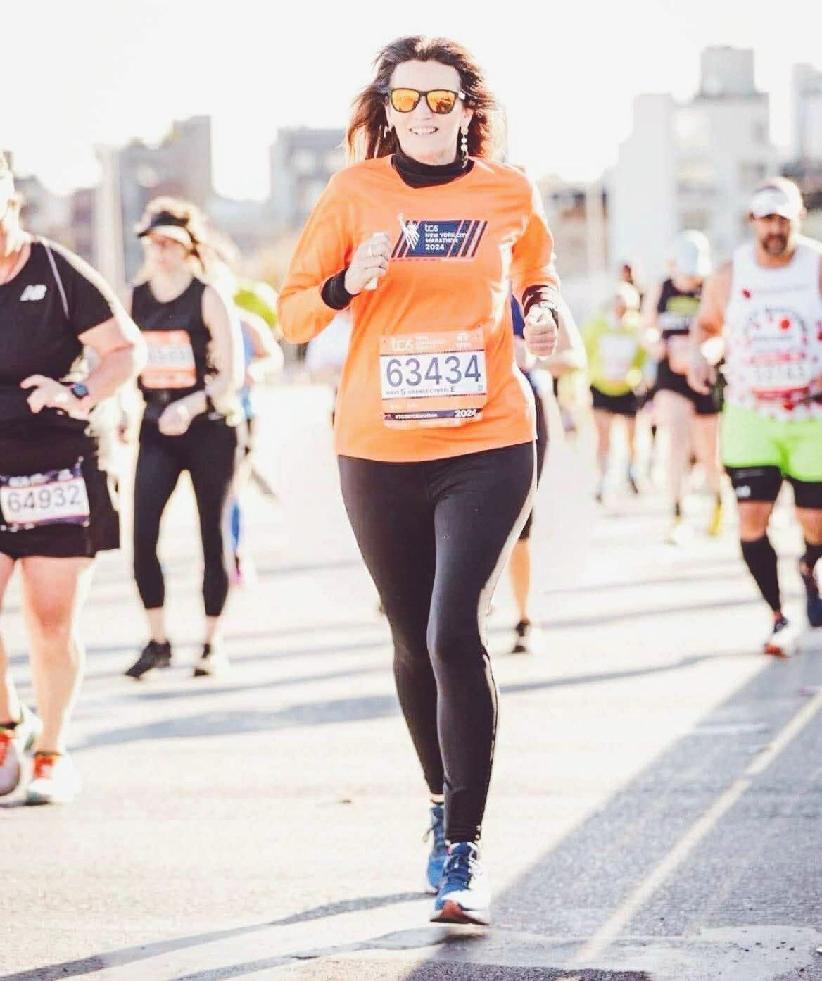 Woman running a marathon, wearing orange shirt and black leggings, with other runners in the background.