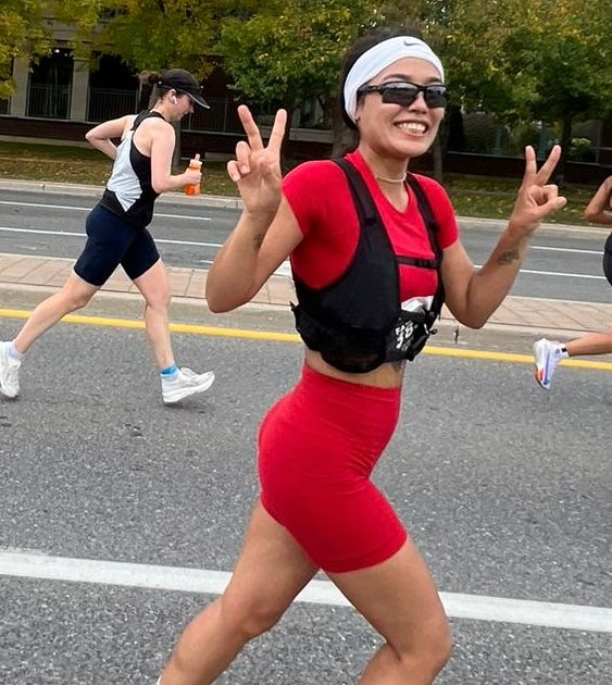 Woman in red running gear giving peace signs during a race. Sunglasses, headband, running vest.