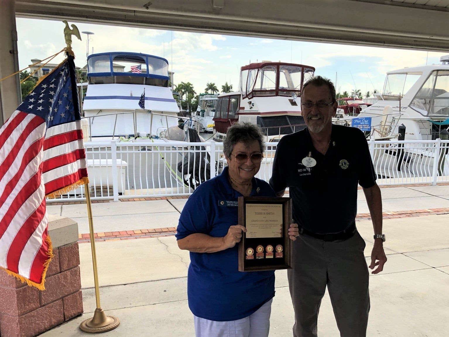Two people holding a framed award near an American flag and boats. Woman in blue and man in black smiling. Outdoor setting.
